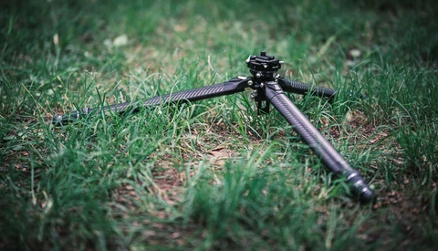 Rifle bipod mounted on grass and soil terrain.