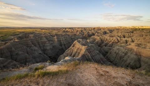 Badlands landscape with eroded buttes, deep ravines, and sparse prairie grass under golden hour light.