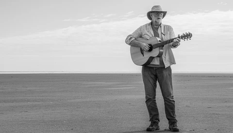 Man playing acoustic guitar while standing alone on a vast, flat desert plain under an overcast sky.