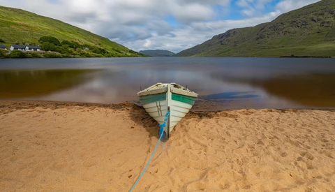 Blue and green rowboat beached on sand with a rope, facing calm water and green mountains under partly cloudy sky.