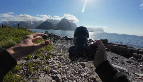Photographer's hands holding a camera on a rocky beach with snow-capped mountains across the water.