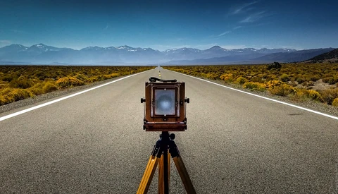Large format camera on tripod centered on a straight desert highway with mountains in the distance.
