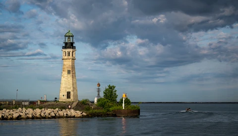 Stone lighthouse with green cupola on rocky breakwater under dramatic cloudy sky.