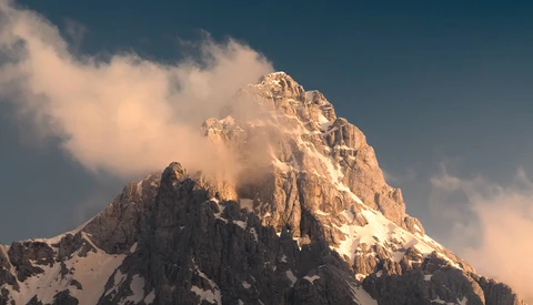 Snow-capped mountain peak bathed in golden light with clouds swirling around its summit.