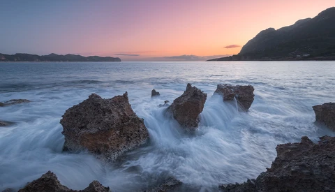 Rocky outcrops surrounded by smooth, flowing seawater at twilight with mountains in the distance.