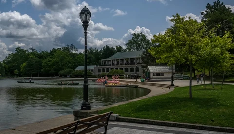Lamppost on wooden dock overlooking a calm lake with historic building and trees under blue sky.