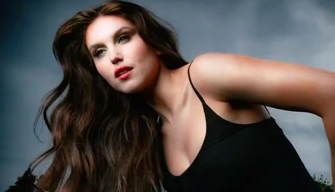 Woman with long dark hair and red lips wearing black tank top against stormy sky background.