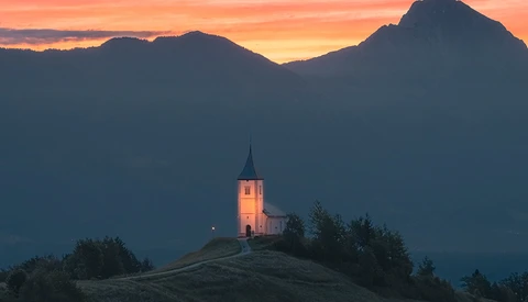 Illuminated church with a steeple on a hilltop at sunset, framed by dark mountains and golden sky.