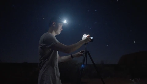 Photographer with headlamp adjusts camera on tripod at night under starry sky.