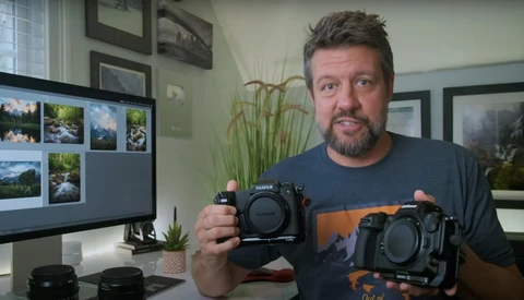 Photographer holding two mirrorless camera bodies side by side in a home office setting.