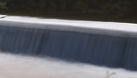 Water flowing over a concrete dam structure with horizontal spillway gates.