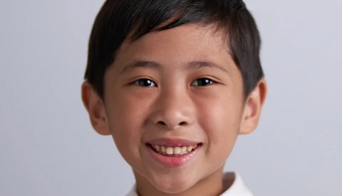 Young boy with dark hair smiling at camera against neutral gray background.