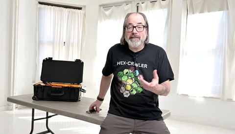 Man wearing glasses and HEX-CRAWLER t-shirt gestures toward a black equipment case on a table in a bright room.