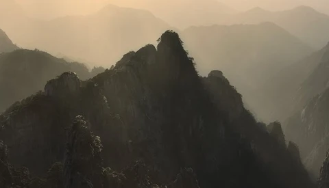 Misty mountain peaks silhouetted against hazy golden light filtering through atmospheric layers.
