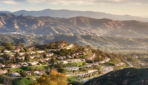Hillside suburban neighborhood with houses nestled among trees, mountains visible in hazy distance.