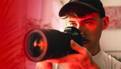 Photographer holding a Nikon camera with a telephoto lens, illuminated by red studio lighting.