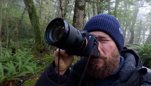 Bearded photographer in dark winter clothing holding a telephoto lens camera in a misty forest.