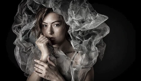Woman with sheer draped fabric around her head and shoulders against a dark background.