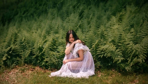 Pregnant woman in white lace dress sitting among tall evergreen trees.