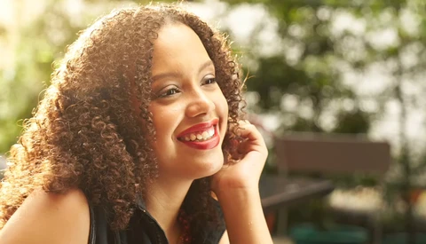 Woman with curly hair smiling outdoors in natural sunlight with blurred foliage background.