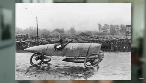 Vintage racing car number 13 on wet track with spectators and trees in background.