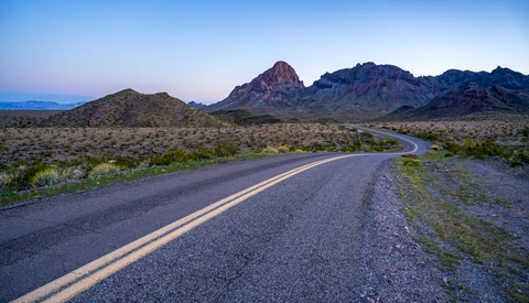 Desert highway with double yellow lines curving toward distant mountains at dusk.