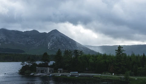 A snow-capped mountain peak rises behind a forested shoreline with a small waterfront cabin and dock.
