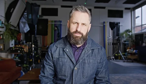 Man with full beard wearing denim jacket in professional studio setting with lighting equipment visible in background.