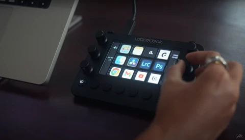 Hand operating a Loupedeck control panel with illuminated customizable buttons and knobs next to a laptop.