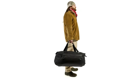 Person in mustard jacket and cap standing against white background while holding black duffel bag.