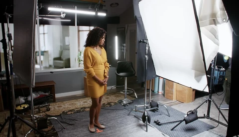 Woman in yellow dress standing in professional photo studio with softbox lighting and reflector setup.