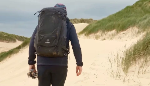 Hiker wearing a large backpack walking away across sand dunes with marram grass.