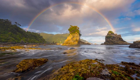 A double rainbow arcs over a dramatic coastal seascape with moss-covered rocks and a prominent sea stack bathed in golden light.