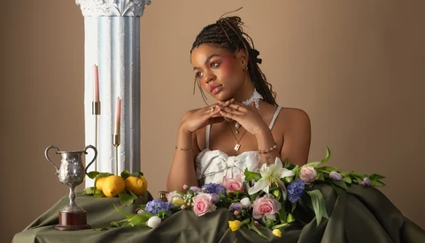 Woman posing with floral arrangement and decorative still life elements against neutral backdrop.
