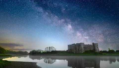 Medieval castle reflected in still water under a starry night sky with the Milky Way visible.