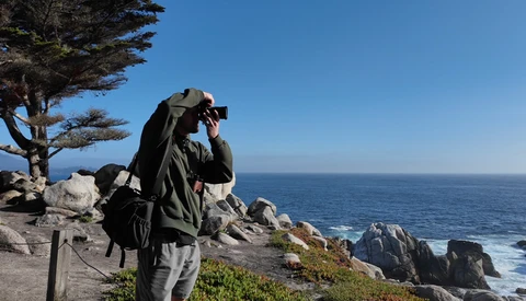 Photographer with camera and backpack on coastal rocks overlooking the ocean.