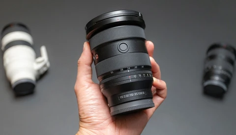 Hand holding a telephoto zoom lens with focus ring and depth-of-field scale visible against gray background.