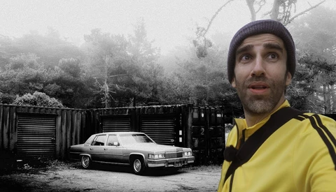 Man in yellow jacket and beanie looking upward beside a vintage car and industrial containers in misty forest setting.