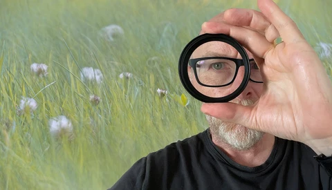 Photographer wearing glasses holds a circular magnifying lens up to his eye, revealing a close-up view of a wildflower meadow behind him.