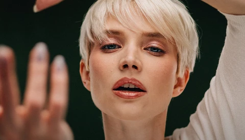 Portrait of a person with platinum blonde pixie cut against a dark background, with hands positioned near their face and shoulder.