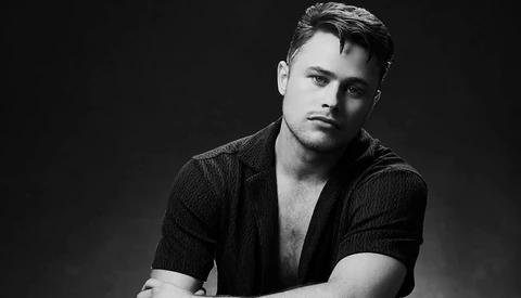 Black and white portrait of a man in an open textured shirt against a dark background.