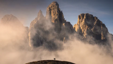 Two hikers standing on a hillside facing towering rock peaks shrouded in mist and clouds.