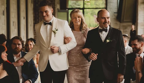 Wedding party walking together down an aisle indoors, with groom in cream blazer, bride in beaded dress, and groomsman in black tuxedo.