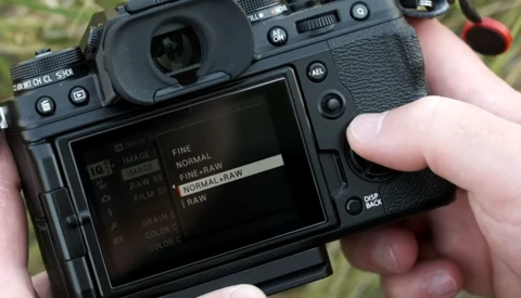 Photographer's hands holding a mirrorless camera with rear LCD screen displaying image quality and file format settings menu.