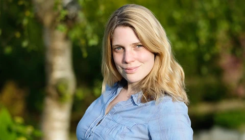 Woman with blonde shoulder-length hair wearing a light blue button-up shirt, photographed outdoors with soft natural lighting.