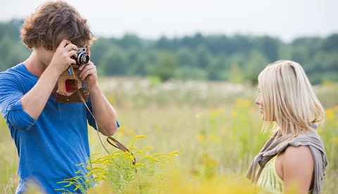 Teenage boy photographing a blonde girl in a wildflower field using a vintage film camera.