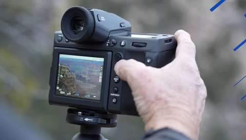 Hand holding a mirrorless camera with articulating LCD screen displaying live view of a landscape scene.