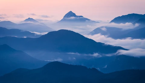 Layered mountain peaks shrouded in mist at dawn, with deep blue tones and a pyramidal summit in the distance.