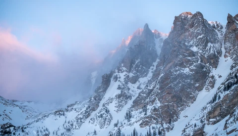 Snow-covered mountain peaks at dawn, with soft pink alpenglow illuminating jagged summits through clearing mist.
