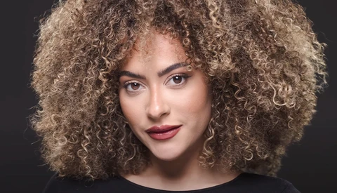 Portrait of a woman with voluminous curly blonde hair against a dark background.